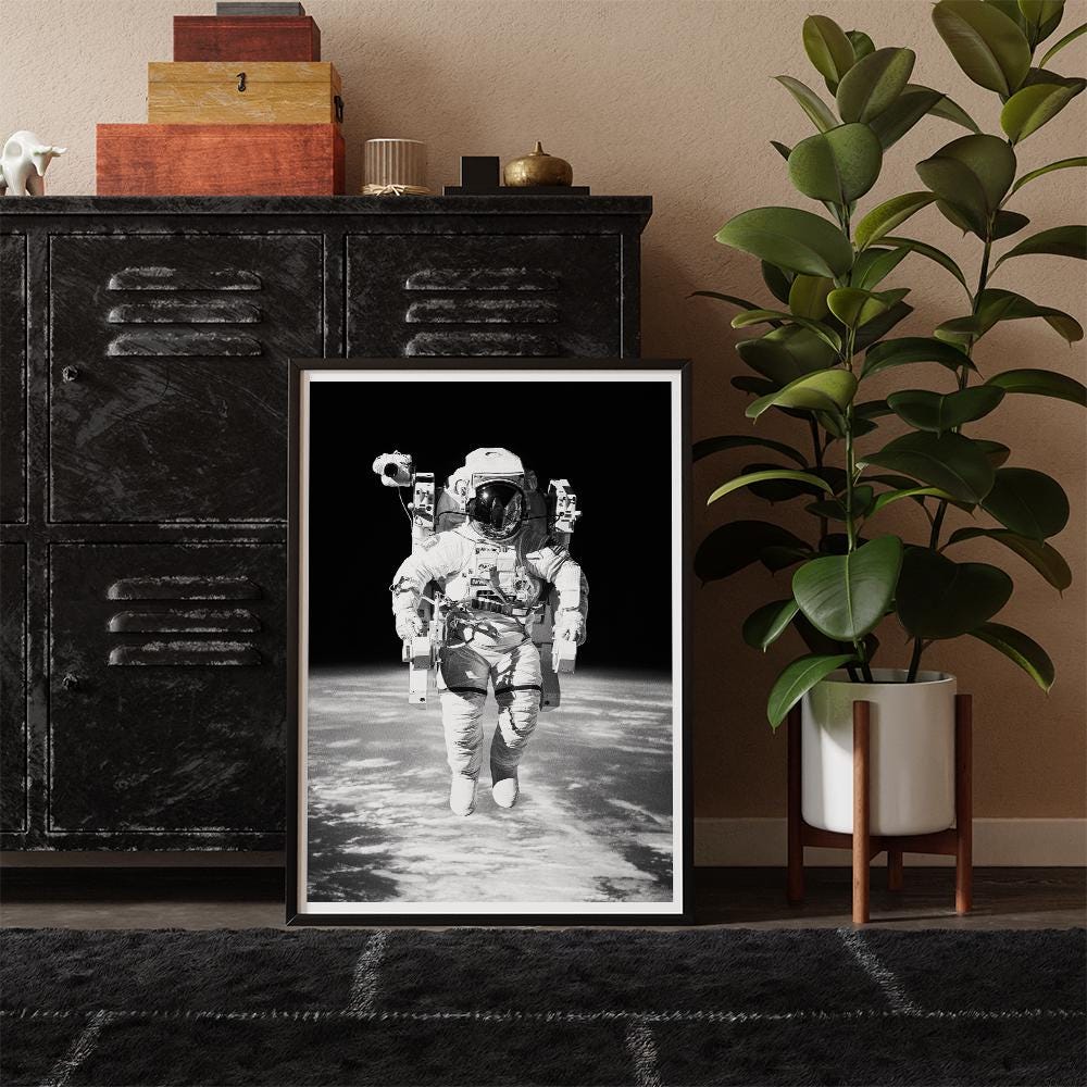 Framed black and white photograph of an astronaut on a floor, with a plant and cabinet in the background.