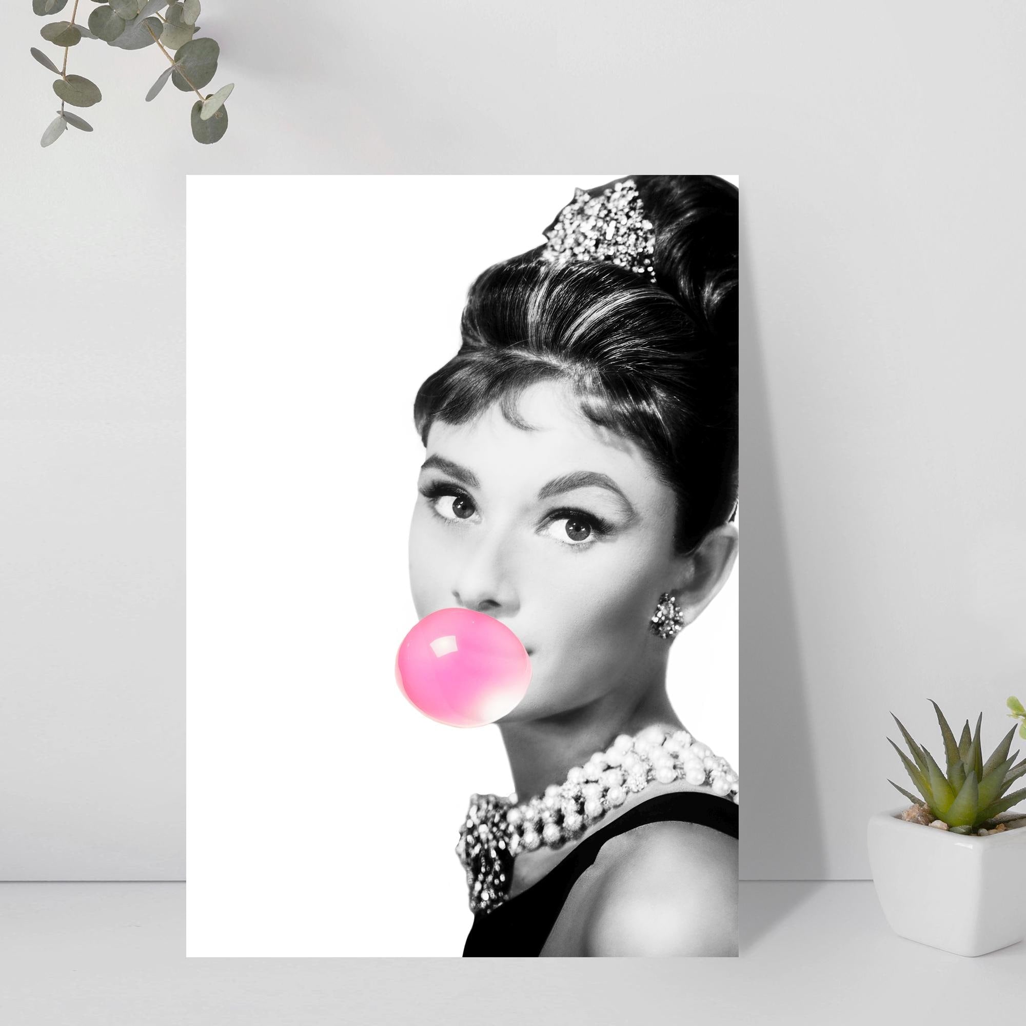 Canvas print of a stylized portrait with Audrey Hepburn blowing a pink bubble gum bubble, displayed on a white wall.
