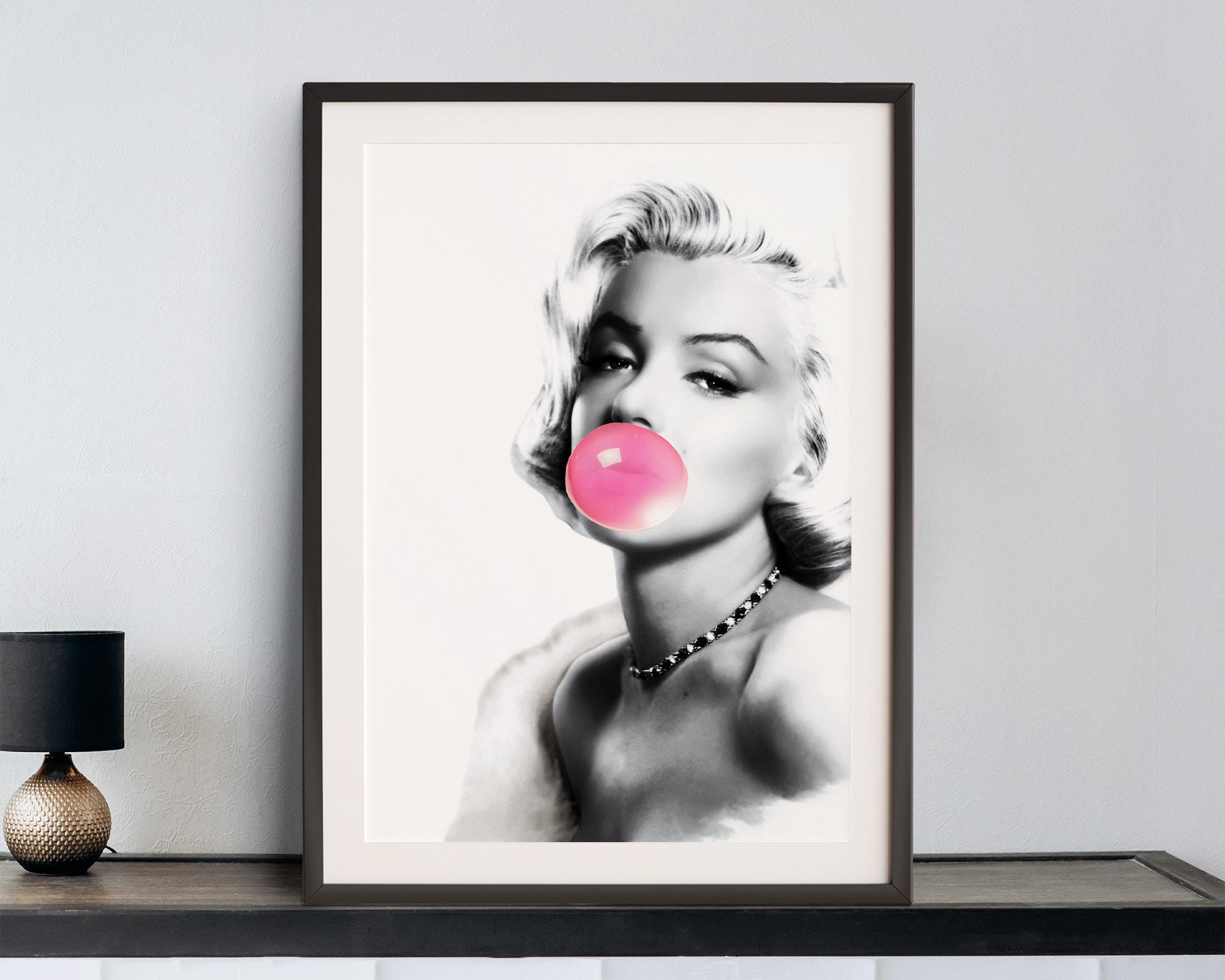 Framed artwork of Marilyn Monroe stylized portrait with a pink bubble gum bubble on a white wall.