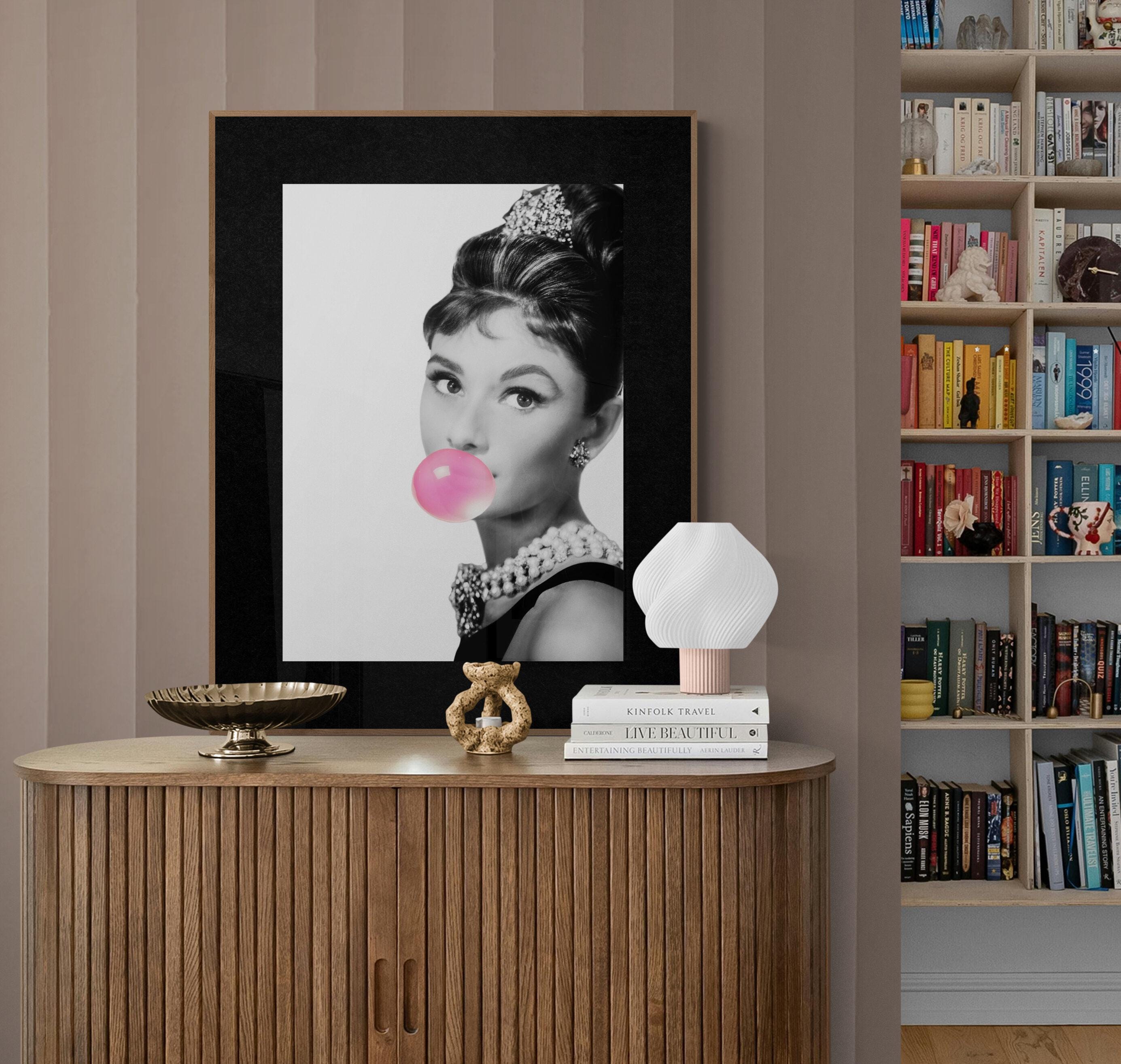 Framed portrait of Audrey Hepburn blowing  a pink bubble gum ball on a wooden side table with decorative items.