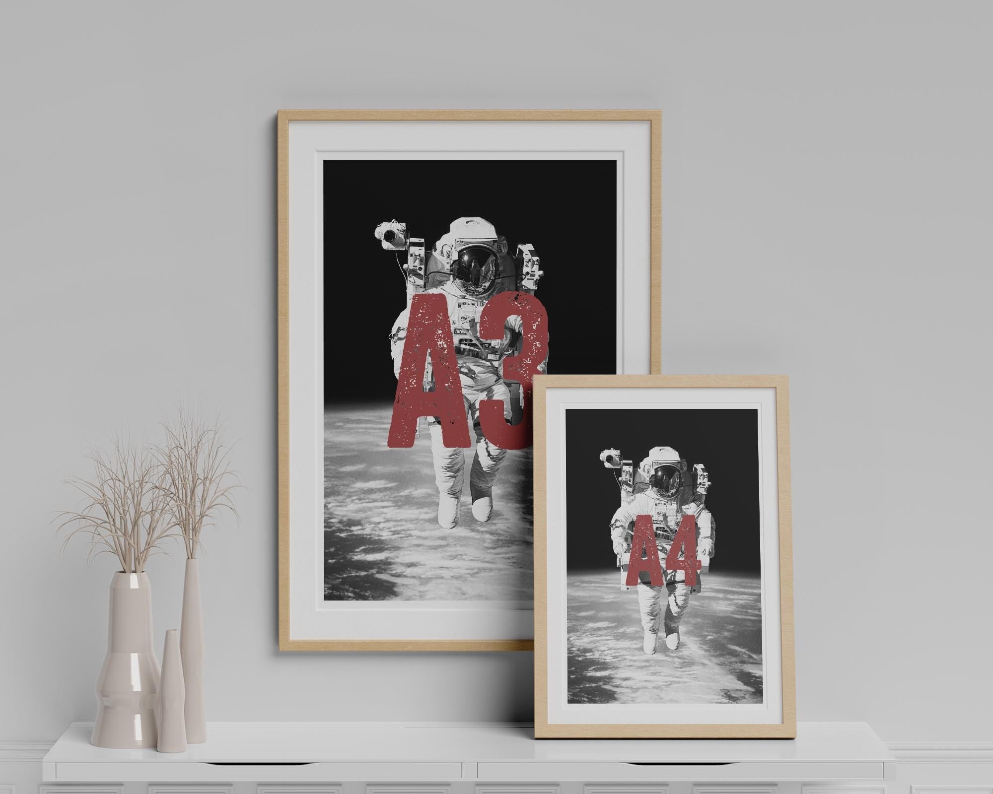 Two framed prints of an astronaut on the moon with 'A3' and 'A4' text, displayed on a shelf against a gray wall.