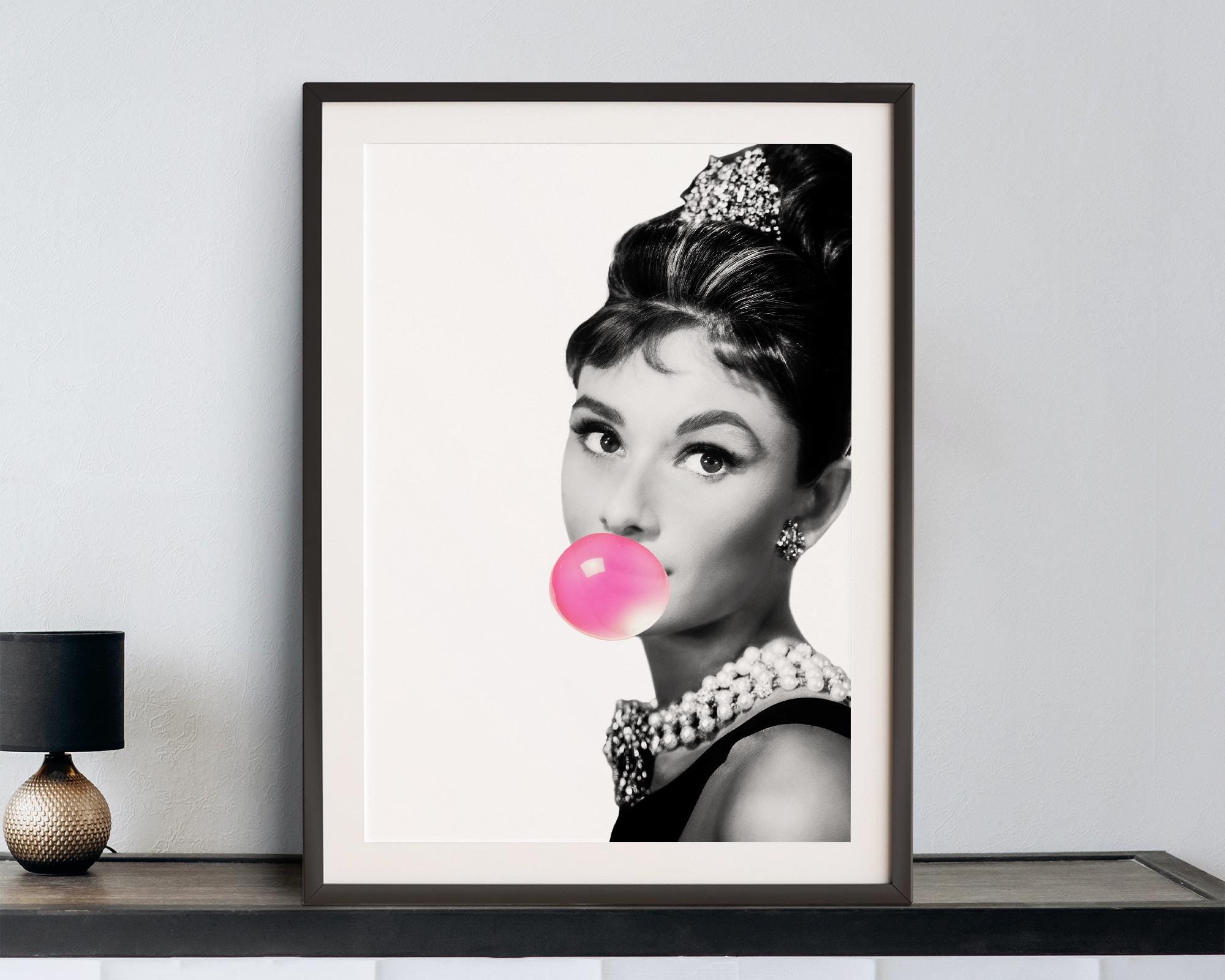 Framed artwork of Audrey Hepburn blowing a pink bubble with gum on a white wall.