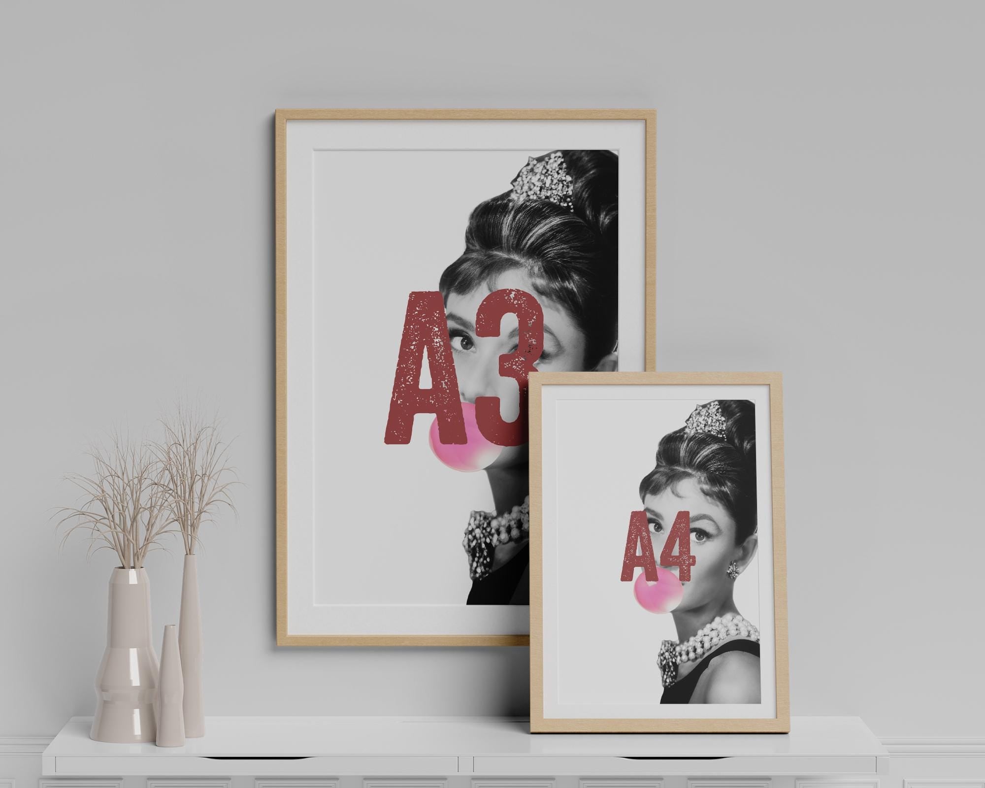 Two framed prints of Audrey Hepburn blowing a bubble with text on a white shelf against a gray wall.