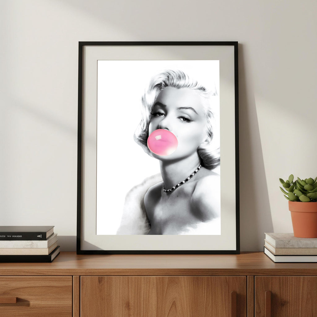 Framed artwork of Marilyn Monroe blowing a pink bubble gum bubble on a wooden surface with books and a plant.