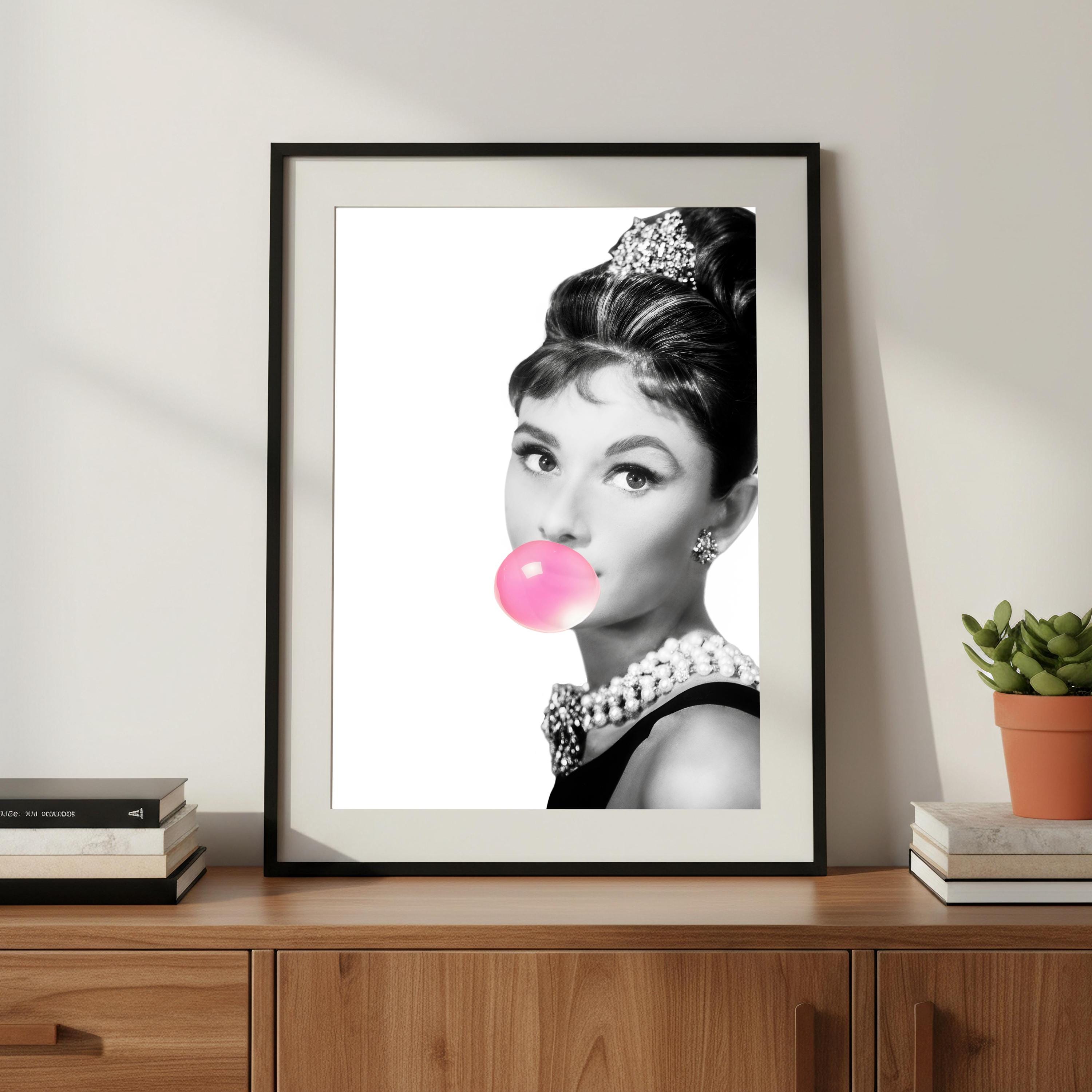 Framed artwork of Audrey Hepburn blowing a pink bubble with a plant and books in the background