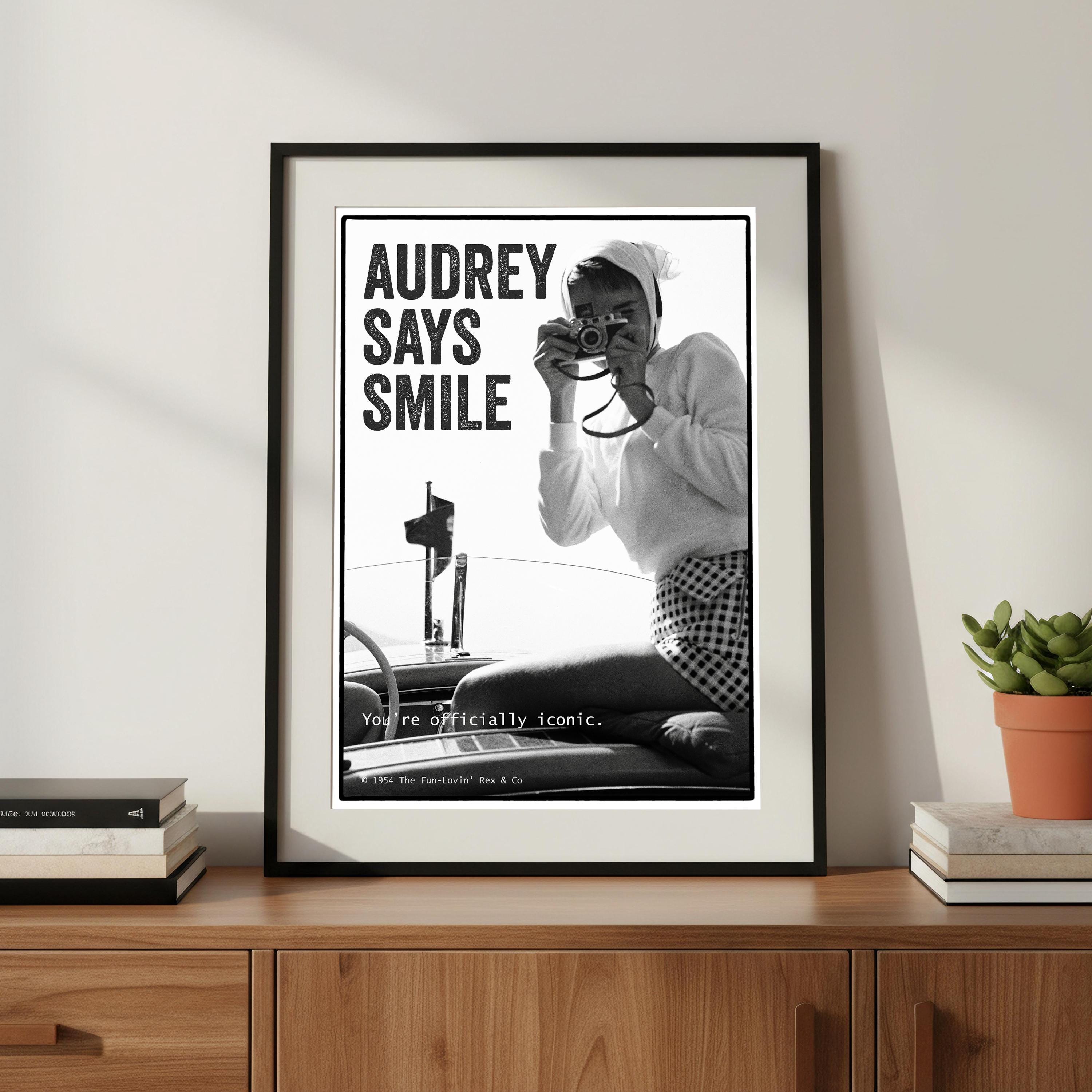 Framed poster with black and white image of Audrey Hepburn taking a photo, text 'AUDREY SAYS SMILE', on a wooden shelf.