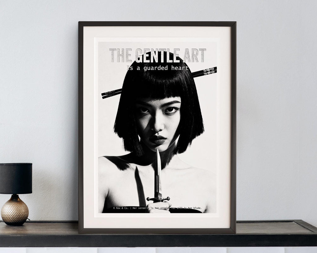 Framed poster with a black and white portrait of a woman with a dagger, text 'The gentle art is a guarded heart', on a light gray wall.