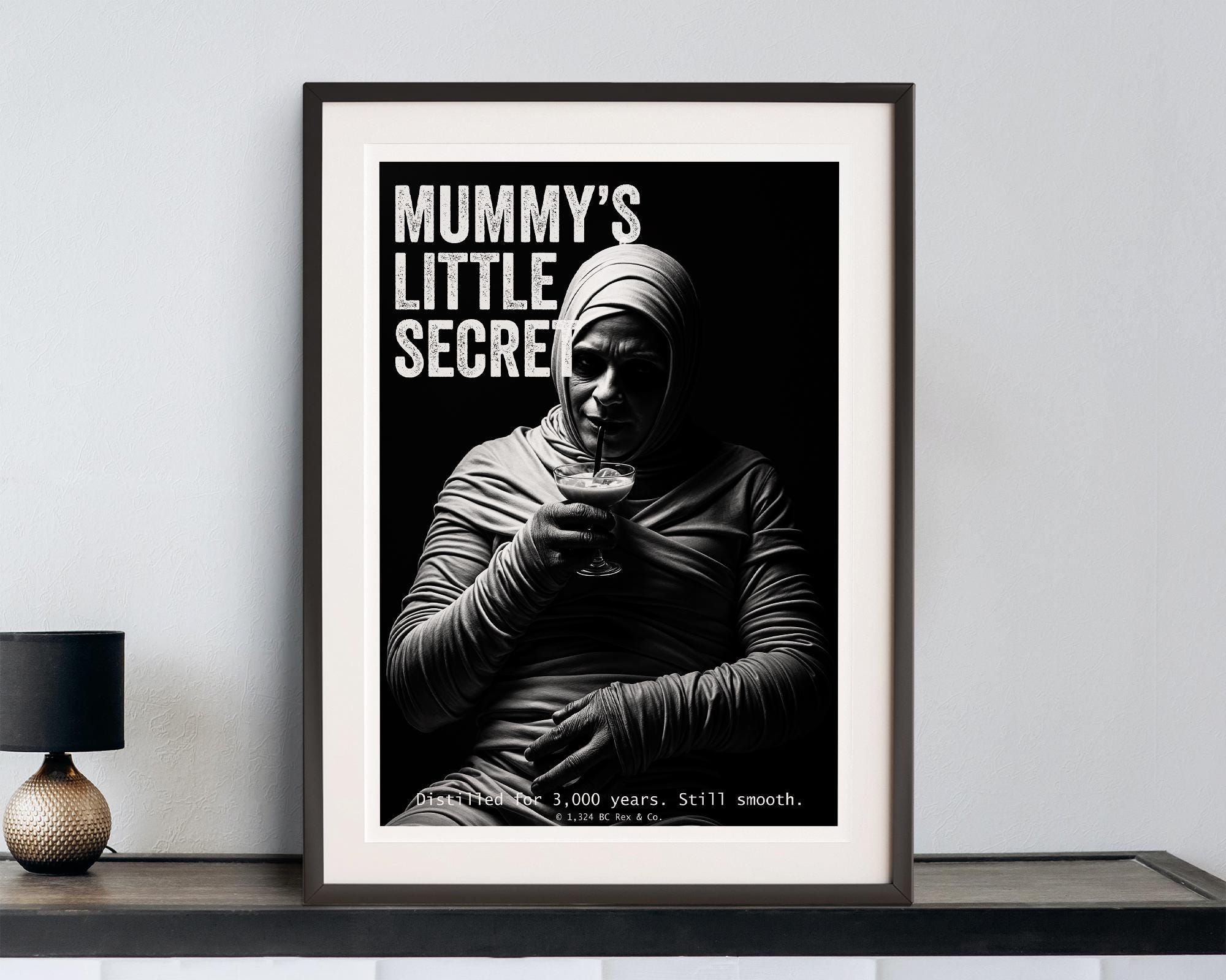 Framed poster with a mummy holding a cocktail and text 'Mummy's Little Secret' on a white wall.
