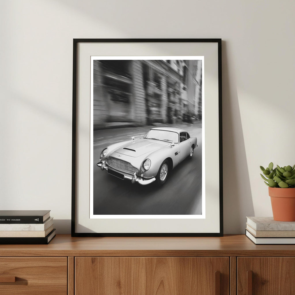 Framed black and white print of a classic car on a wooden shelf with books and a plant.