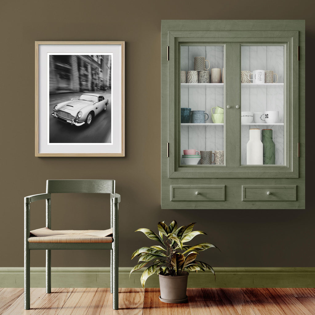 Framed picture of an Aston Martin DB5 next to a Green cabinet with glass doors, chair, and plant against a brown wall.