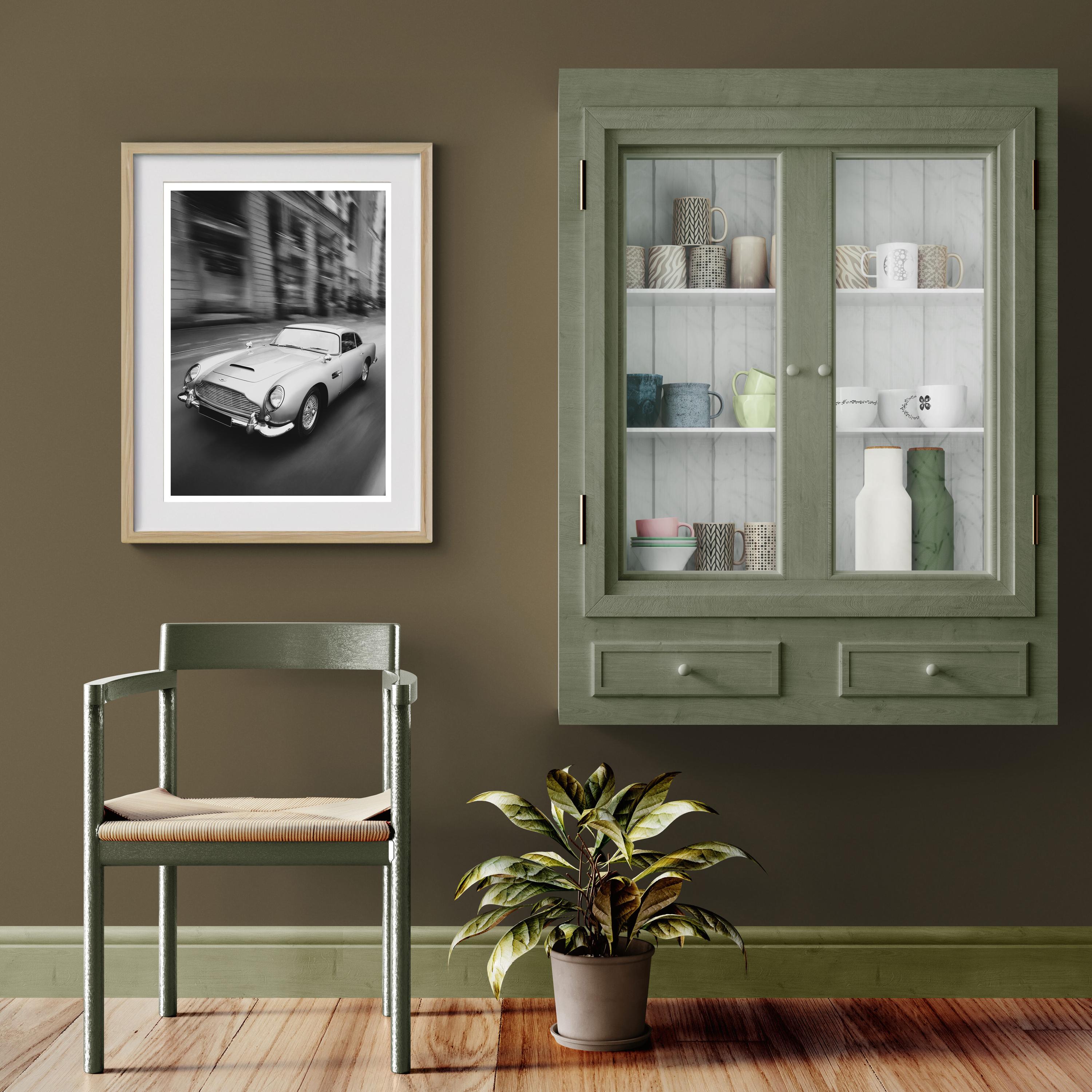 Framed picture of an Aston Martin DB5 next to a Green cabinet with glass doors, chair, and plant against a brown wall.