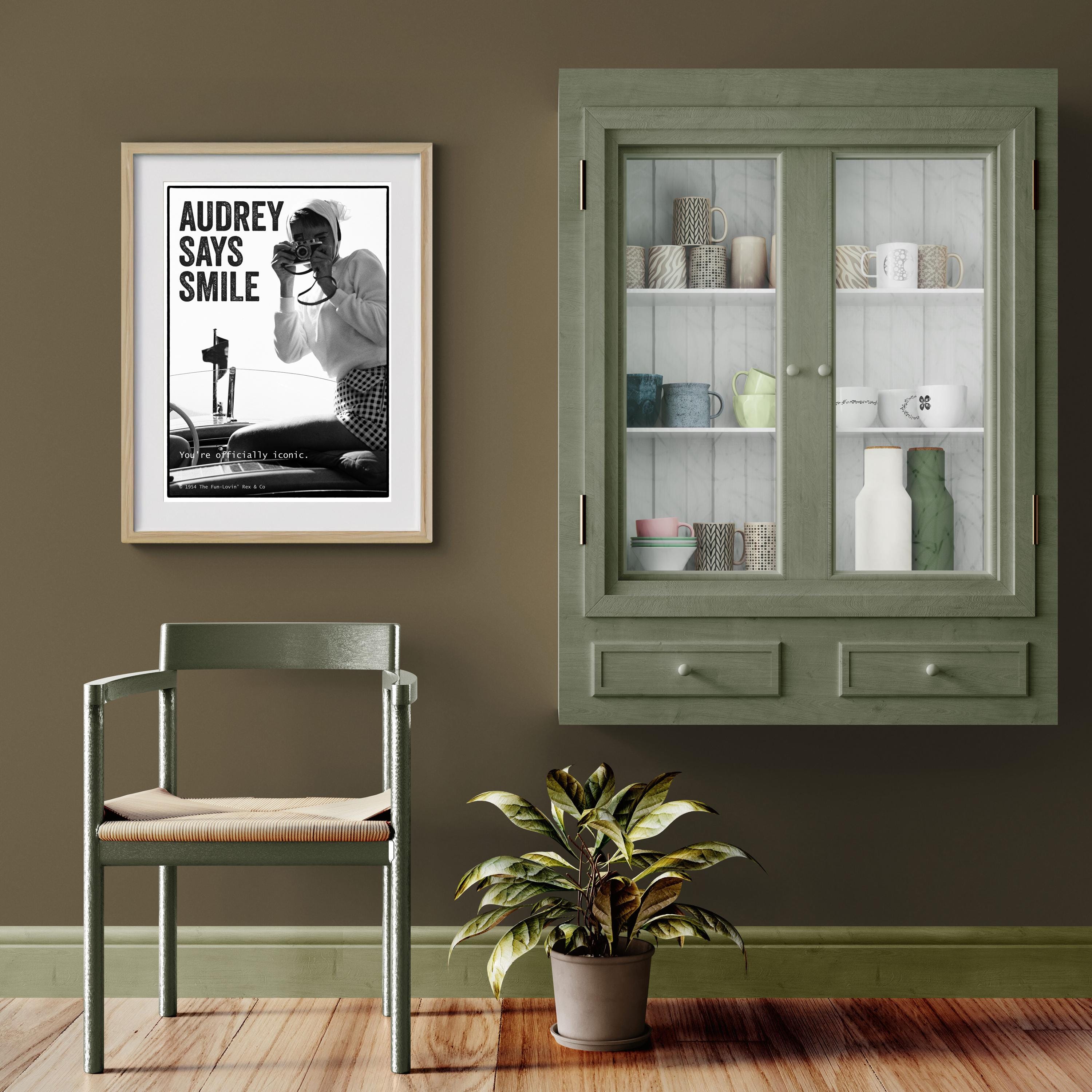 Room interior with a framed poster of Audrey Hepburn, green cabinet, chair, and plant on a wooden floor.